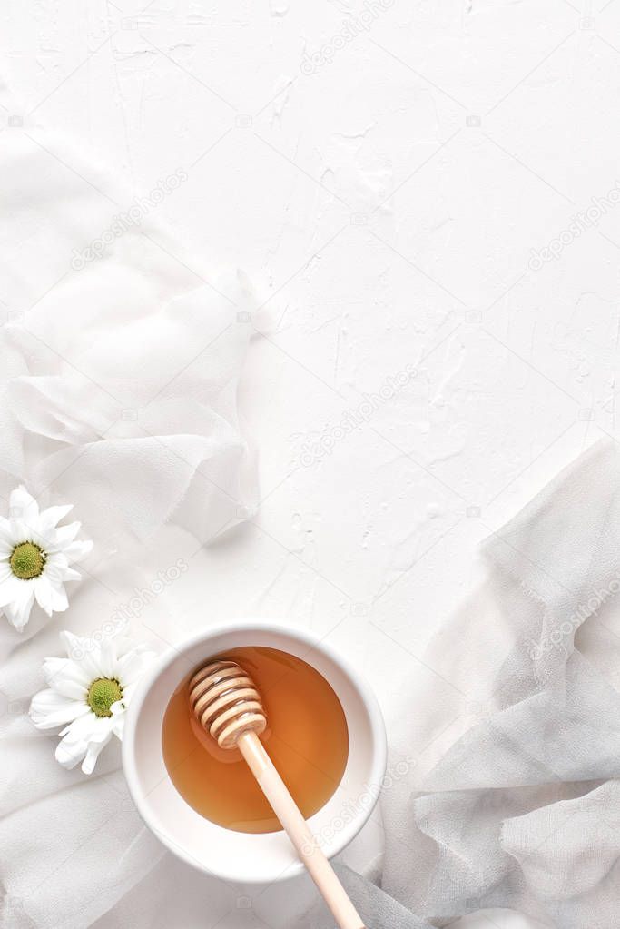 Flat lay of honey in small ceramic bowl, wooden honey dipper, flowers, petals on a white background with copy space. Top view.