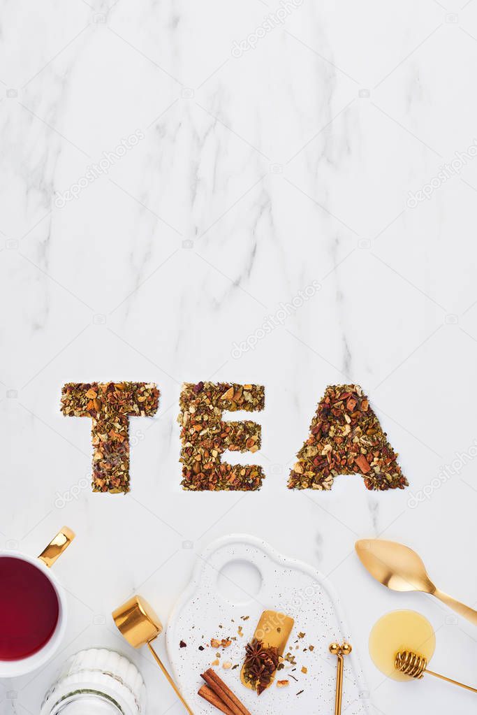 Tea spelled out with dried herbs and fruits on white marble background with tea utensils at the bottom. Flat lay, copy space. Tea concept, top view.