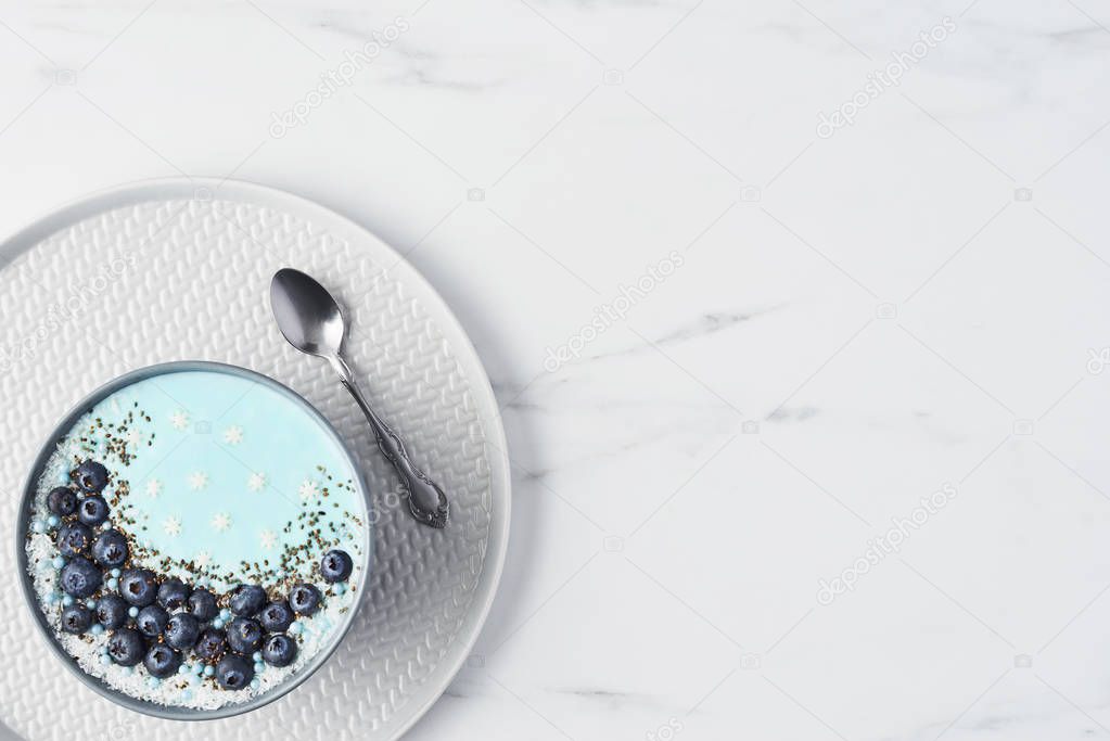 Top view of blue yogurt smoothie bowl made with blueberry, coconut flour, chia seeds and sugar pearls on white marble table with copy space.