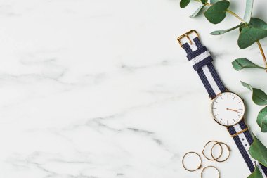 Women's watch with navy blue and white nylon strap, golden rings and eucalyptus on white marble background. Top view with copy space for text. clipart