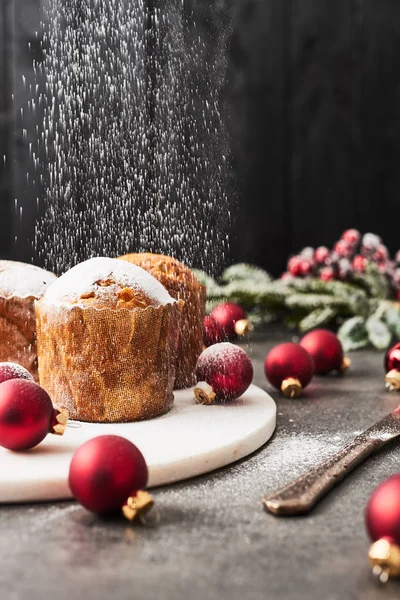 将糖粉洒在传统的圣诞迷你 Panettone 上面有葡萄干和干果 在白色大理石上 盘子里环绕着黑色木制背景的混凝土桌子上的红色小泡 — 图库照片
