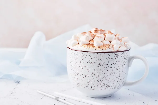 Hot chocolate or cocoa with whipped cream and marshmallow candy sprinkled with cinnamon or cocoa powder with straws on white marble table over blue silk and pink background with copy space for text.