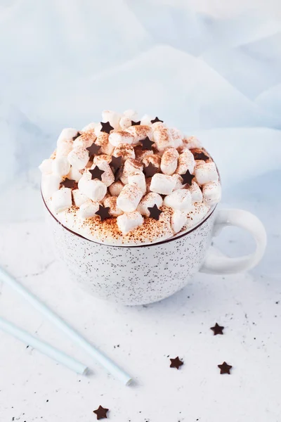 Hot chocolate or cocoa with whipped cream and marshmallow candy sprinkled with cinnamon or cocoa powder with chocolate stars on white marble table and blue silk in the background with copy space.
