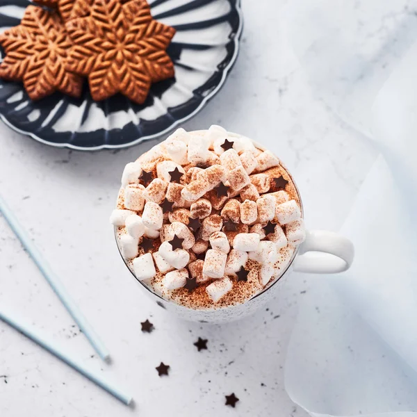Hot chocolate or cocoa with whipped cream and marshmallow candy sprinkled with cinnamon or cocoa powder with chocolate stars and Christmas gingerbread cookies on blue serving plate. Square crop.