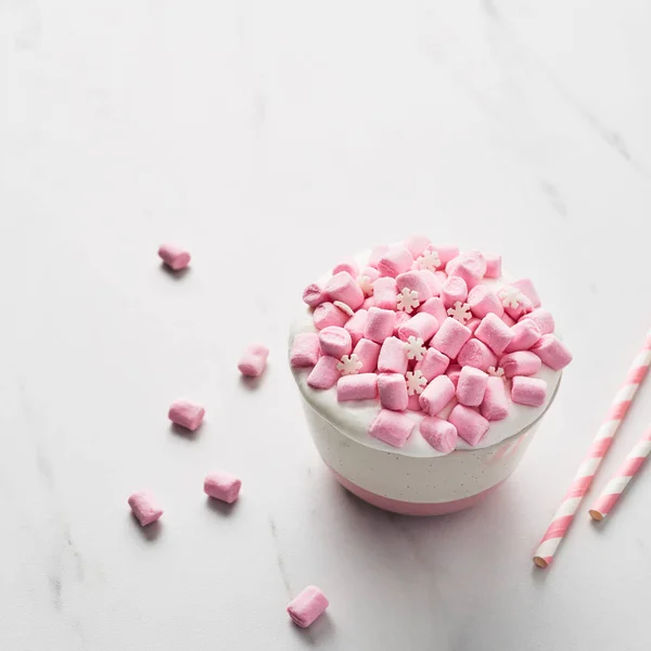 Hot chocolate or cocoa with whipped cream and pink marshmallow candy in pink and white mug with pink striped straws on white marble table. Copy space for text. Square crop.