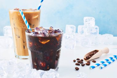 Yaz içeceği bir bardak kahve ve buzlu kahve krem uzun bir bardak, kahve çekirdekleri ve mavi arka plan üzerinde beyaz mermer masa buza çevrili payet ile buzlu. Metin için yer kopyalayın.