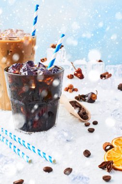 Kış içecek bir bardak kahve ve buzlu kahve krem buz, kahve çekirdekleri ve beyaz mermer tablosundaki çeşitli baharatlar kar ile mavi arka plan üzerinde çevrili uzun bir bardak buzlu. Kopya alanı.