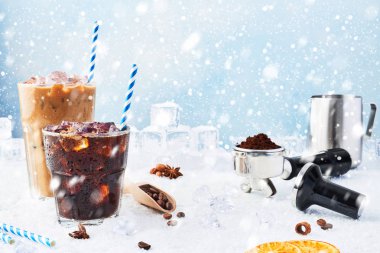Winter drink iced coffee in a glass and ice coffee with cream in a tall glass surrounded by ice, coffee beans, portafilter, tamper, milk jug and various spices on snow over blue background. Copy space clipart