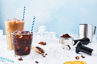 Yaz içeceği buzlu cam ve buz kahve kahve krem buz, kahve çekirdekleri, portafilter, sabotaj, süt kabı ve çeşitli baharatlar kar üzerinde mavi arka plan üzerinde çevrili uzun bir bardak. Kopya alanı