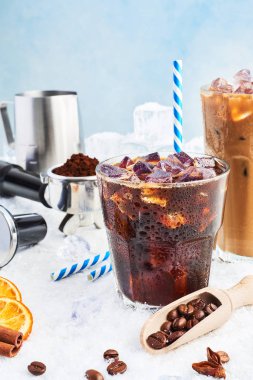 Cam ve buz kahve krem buz, kahve çekirdekleri, portafilter, sabotaj, süt kabı ve çeşitli baharatlar kar üzerinde mavi arka plan üzerinde çevrili uzun bir bardak buzlu içecek kahve veya soda yaz.