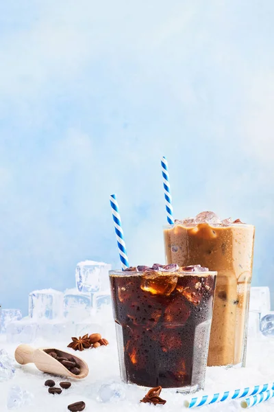 Summer drink iced coffee or soda in a glass and ice coffee with cream in a tall glass surrounded by ice cubes, coffee beans and various spices on snow over blue background. Copy space for text.
