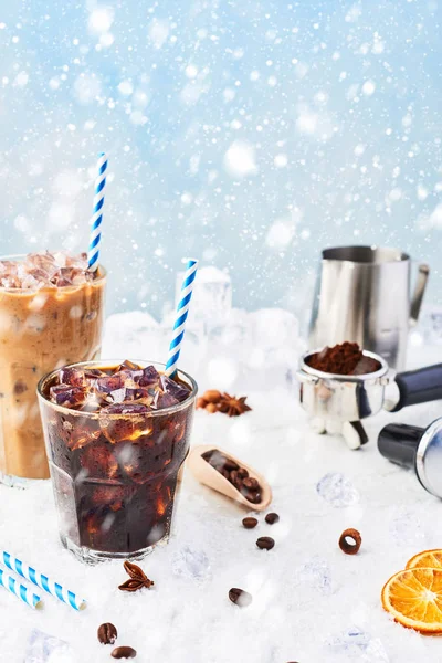 Winter drink iced coffee in a glass and ice coffee with cream in a tall glass surrounded by ice, coffee beans, portafilter, tamper, milk jug and various spices on snow over blue background. Copy space