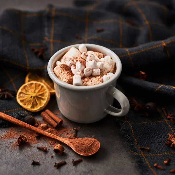 Hot cocoa drink with marshmallows in retro mug surrounded by ingredients: cinnamon, orange, anise and cove on black concrete table. Winter drink. Top view. Rustic look. Copy space. Square crop.
