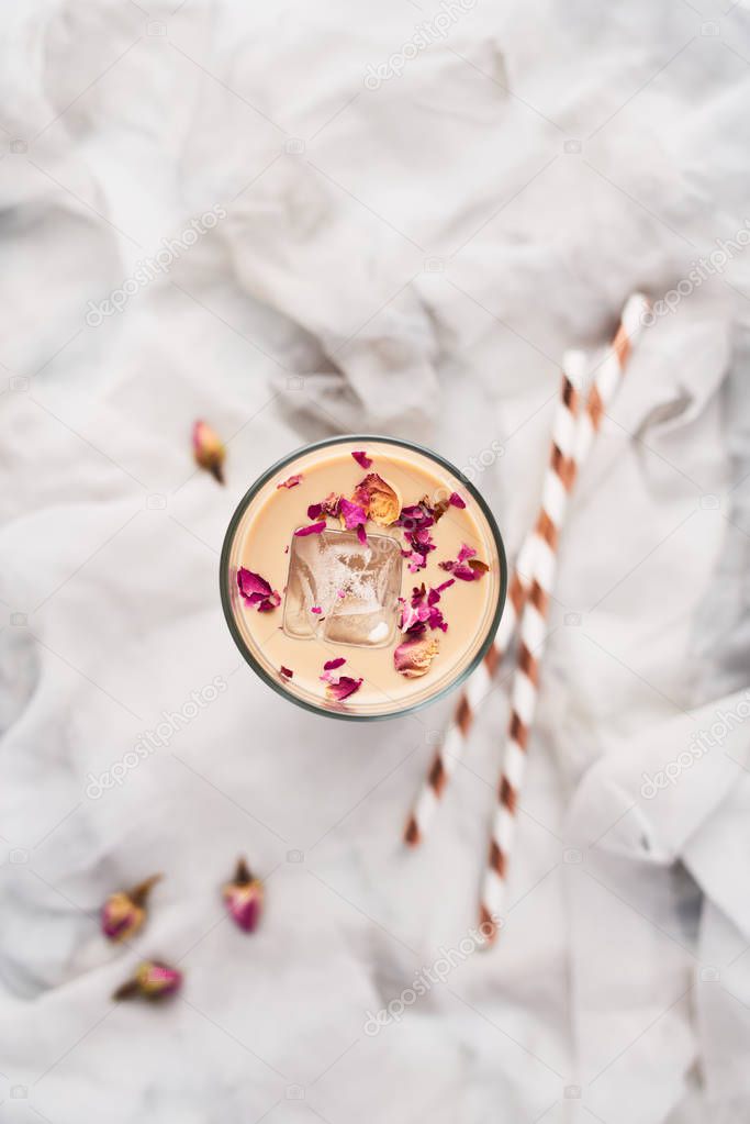 Iced coffee with rose and cardamom in a tall glass surrounded by dried roses and straws on white and gray silk background. Top view with copy space for text.