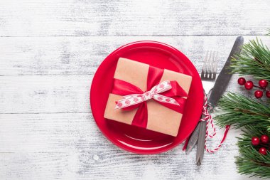 Kırmızı tabaklı Noel masası seti, hediye kutusu ve hafif ahşap arka planda gümüş takımlar. Ağaç dalları, kutsal meyveler. Kopyalama alanı olan üst görünüm.