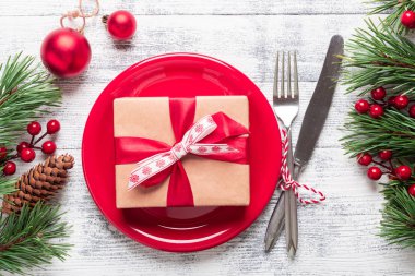 Kırmızı tabaklı Noel masası seti, hediye kutusu ve hafif ahşap arka planda gümüş takımlar. Ağaç dalları, kutsal meyveler. Kopyalama alanı olan üst görünüm.