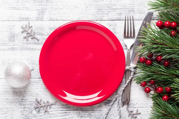 stock image Christmas table setting with empty red plate, gift box and silverware on light wood background. Fir tree branch, holly berries. Top view with copy space.