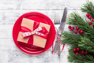 Kırmızı tabaklı Noel masası seti, hediye kutusu ve hafif ahşap arka planda gümüş takımlar. Ağaç dalları, kutsal meyveler. Kopyalama alanı olan üst görünüm.