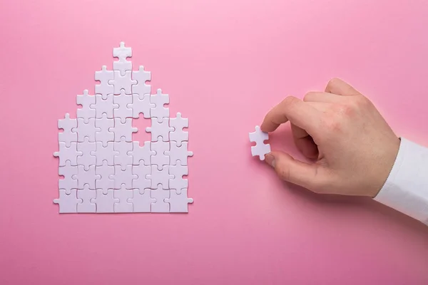 Beyaz bulmaca. Ev şekli bulmaca. Kavram kira, ev kredisi. Beyaz bulmacanın parçası tutan el. Pembe bir arka plan.
