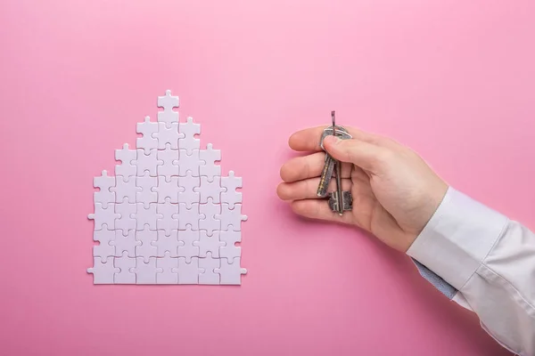 White puzzle. House shape puzzle. The concept of rent, mortgage. Hand holding keys. Pink background. Top view Top view. Copy space