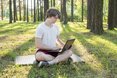 Genç adam dizüstü bilgisayarıyla parkta çimlerin üzerinde oturuyor. Uzaktan öğrenme konsepti. - Görüntü