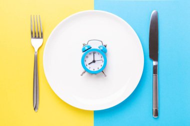 Blue alarm clock, fork, knife and empty plate on colored paper background. Intermittent fasting concept - Image clipart