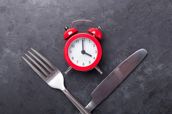 Red alarm clock, fork, knife and empty black ceramic plate on dark stone background. Intermittent fasting concept - Image