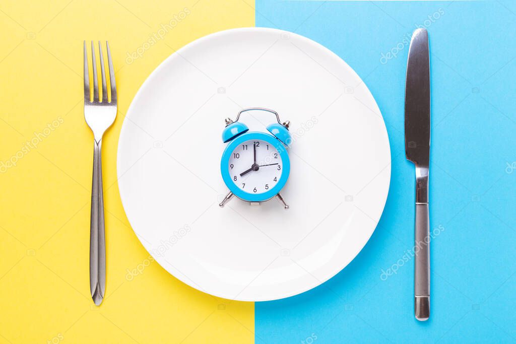 Blue alarm clock, fork, knife and empty plate on colored paper background. Intermittent fasting concept - Image