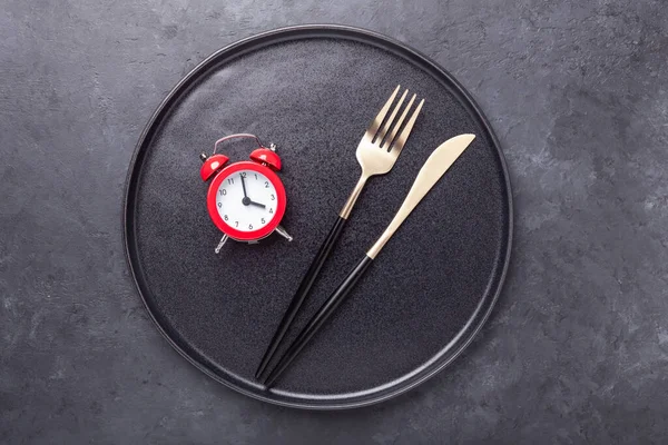 Red alarm clock, fork, knife and empty black ceramic plate on dark stone background. Intermittent fasting concept - Image