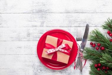 Kırmızı tabaklı Noel masası seti, hediye kutusu ve hafif ahşap arka planda gümüş takımlar. Ağaç dalları, kutsal meyveler. Kopyalama alanı olan üst görünüm.