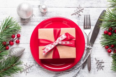 Kırmızı tabaklı Noel masası seti, hediye kutusu ve hafif ahşap arka planda gümüş takımlar. Ağaç dalları, kutsal meyveler. Kopyalama alanı olan üst görünüm.