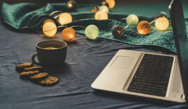Tea with Lemon and Cookies with Chocolate Laying on Mattress - Christmas Decoration Light Balls and Grey Laptop Besides it, Vintage Look Edit