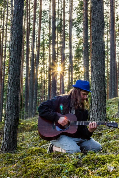 Young and Handsome Guy Playing Guitar in Forest with a Blue Hat on his Head on a Sunny Early Spring Day - Moody Photo with a Sun Star Shining over his Shoulder with a little Sun Flare