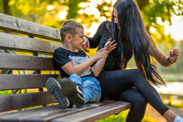 Mom Tickles Her Son on a Park Benck  in Autum with Colorful Backgroun in a Sunny Day, Both Laughing- Caption on Shirt \