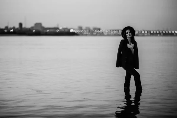 Una chica atractiva en un traje y sombrero se encuentra en el agua sobre el fondo de la ciudad — Foto de Stock