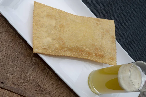 Brazilian food: pastel, or pastry in English, on white dish, and glass of sugar cane juice, on rustic wooden table