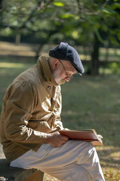 Old man with a white beard sitting on the park bench reading a book, with trees in the background. Brazil