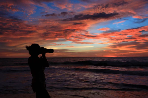 Silhouette of young woman photographer on seashore against beautiful sunset