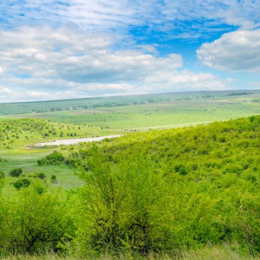 Nehri Vadisi tepeler ve teraslar ile. Yamaçlarda çalılar, ağaçlar ve otlar büyür. Cumulus bulutları mavi gökyüzünde ışık.