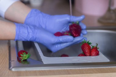 Mavi lateks eldivenli eller, meyveleri koronavirüsten arındırmak için çilekleri dezenfekte ediyor. Mutfak lavabosunda meyveyi suyla ve kül suyuyla yıkıyorum..