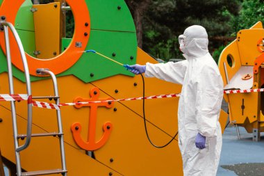 Çocuk parkı virüslere ve hastalıklara karşı dezenfekte ve sterilize ediyor. Beyaz koruyucu giysili bir adam durmak için dezenfektan sıkıyor.