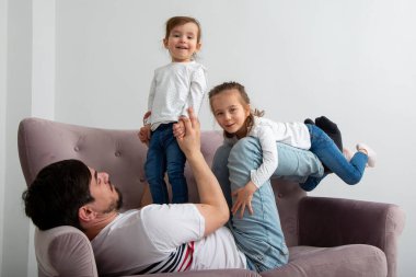 Mutlu bir aile. Genç baba, küçük tatlı kızı kaldırıyor uçak gibi uçuyor güneşli bir haftasonu kanepesinde eğleniyor, kaygısız, sevimli küçük çocuklar, anne babasıyla kanepede oynuyor.