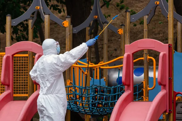 Çocuk parkı virüslere ve hastalıklara karşı dezenfekte ve sterilize ediyor. Beyaz koruyucu giysili bir adam durmak için dezenfektan sıkıyor.