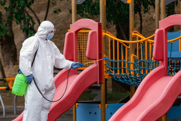 Çocuk parkı virüslere ve hastalıklara karşı dezenfekte ve sterilize ediyor. Beyaz koruyucu giysili bir adam durmak için dezenfektan sıkıyor.