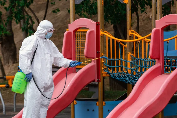 子供たちは ウイルスや病気を消毒して消毒する遊び場 スライド上で消毒剤を噴霧白い保護スーツの男が停止する ロイヤリティフリーのストック画像
