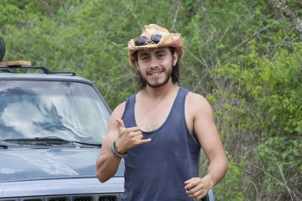 Handsome male Hispanic adventurer smiles and makes hang loose sign with hand in front of truck parked in wilderness