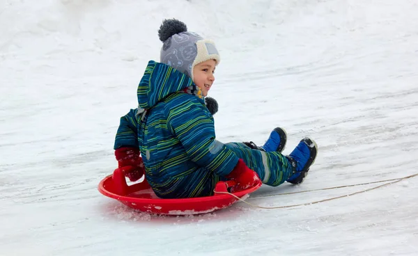 一个4岁的孩子 一个男孩 骑着雪橇从雪滑梯上走 他穿着冬衣 面带微笑 — 图库照片