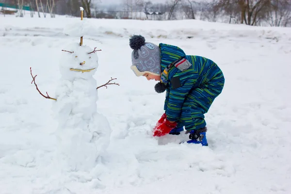 少年は 笑みを浮かべてロール雪の玉と雪だるまを Sculpts します 近くには 小さな雪だるまです — ストック写真