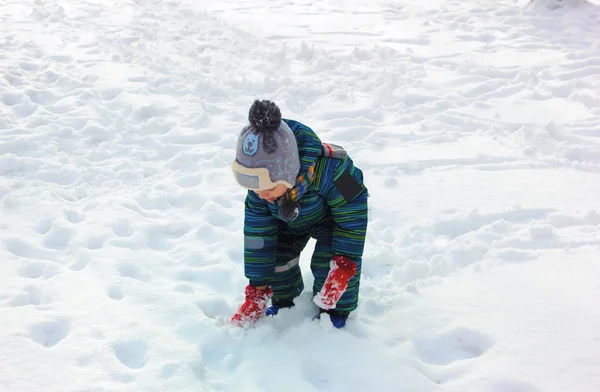 カラフルな冬の服を着た男の子は晴れた冬の日に雪に覆われた地面に雪で遊ぶ — ストック写真