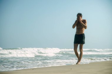 Bir adam Yüzme şort büyük dalgalar sörf yanında elleriyle yüzünü kaplayan kumlu bir plaj boyunca yürüyor. Açık gökyüzü, güneşli bir gün.